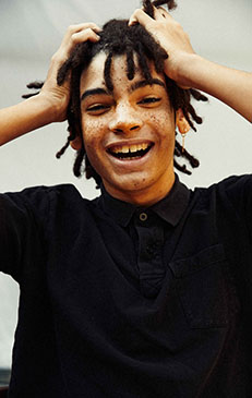Portrait of a Black teenage boy with freckles and dreadlocks posing indoors, wearing a black polo shirt and looking at the camera.