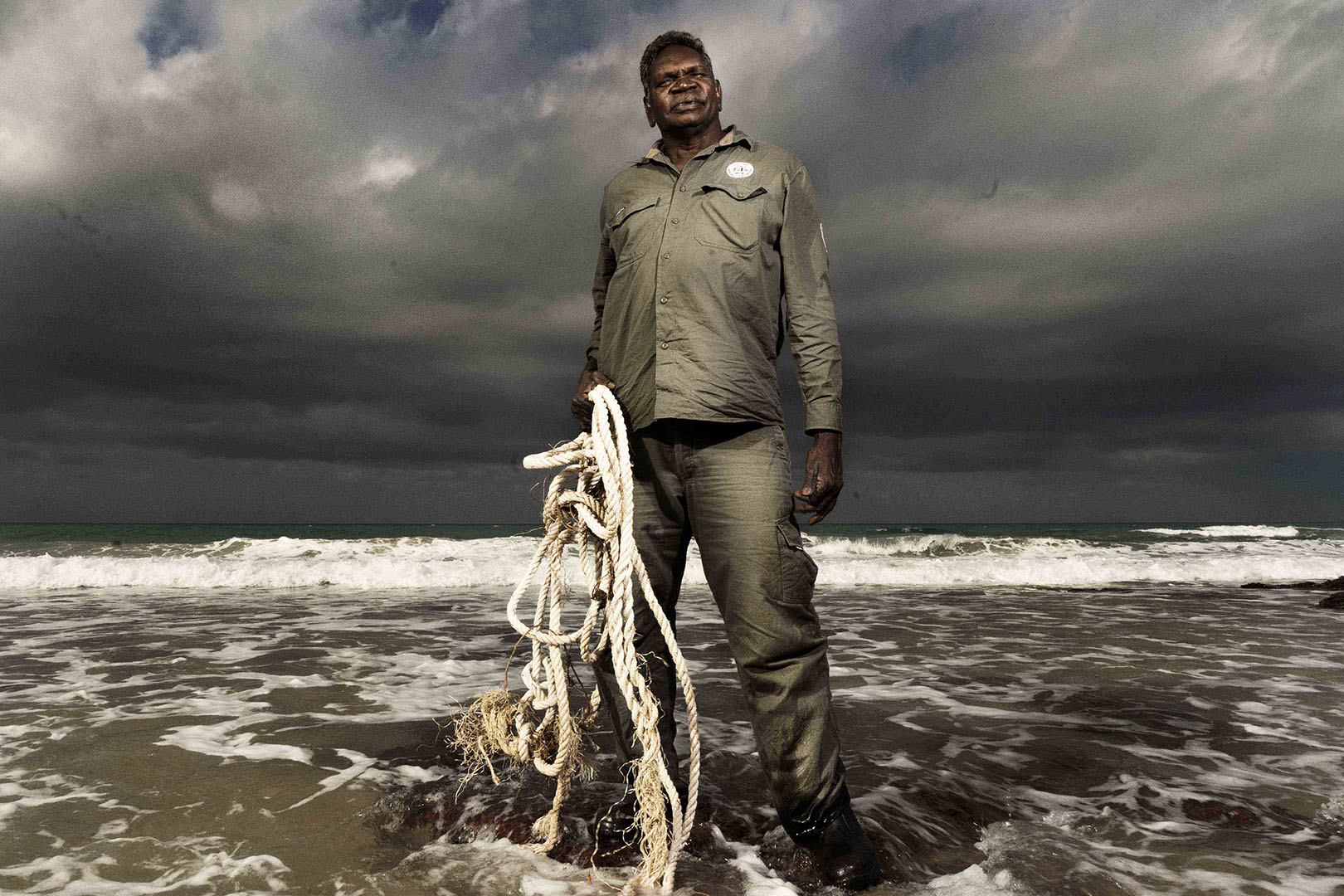 Adult man stands on a beach at the water's edge holding a tangled rope. Stormy sky and waves are visible in the background.