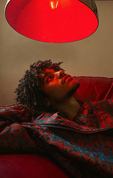 A young Black man with dreadlocks reclines on a red couch under a glowing red lamp, wearing a patterned robe in an indoor setting.