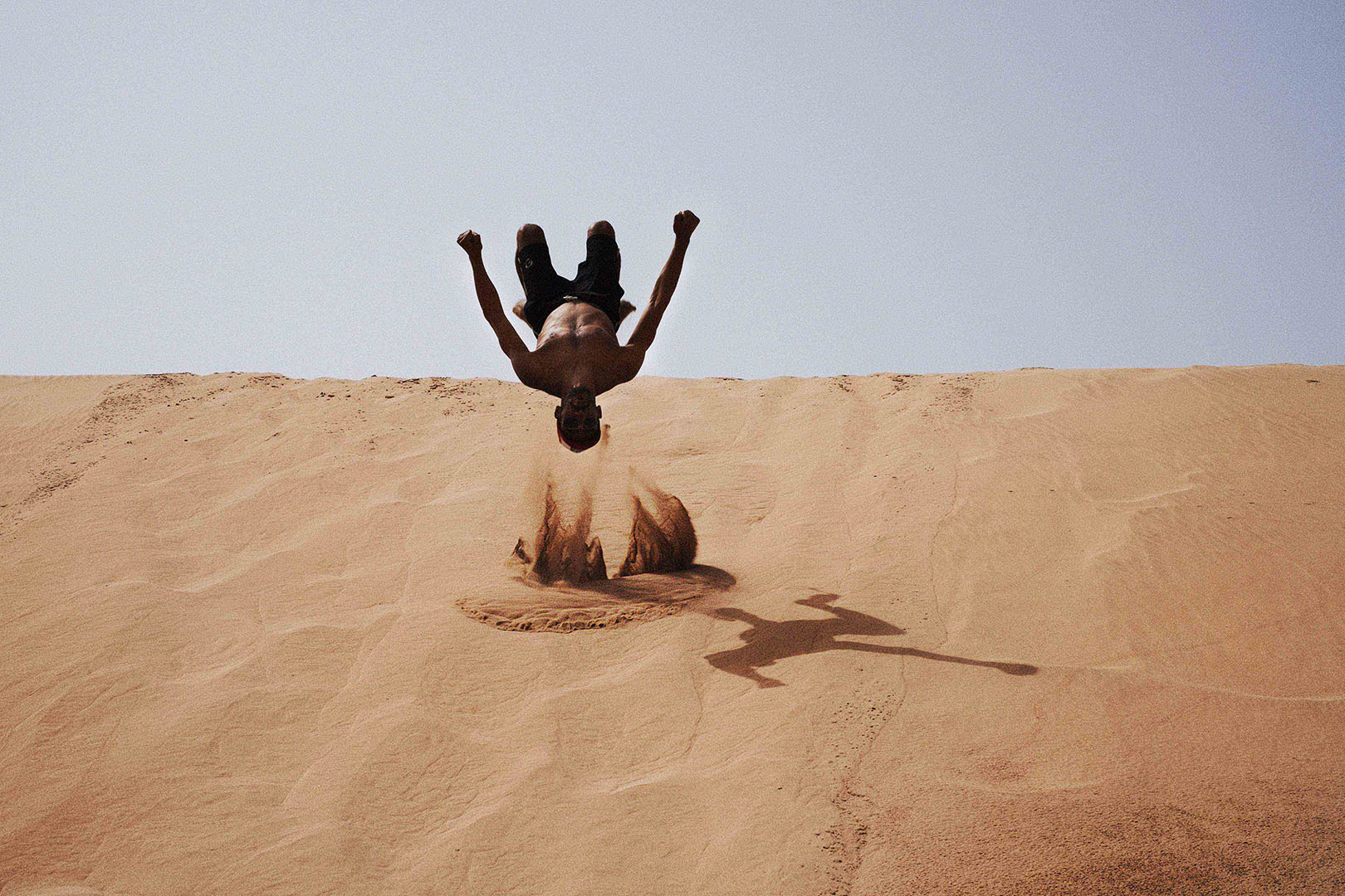 Athletic shirtless man in mid-air performing a backflip on a tan sand dune against a clear sky, showcasing fitness and outdoor activity.