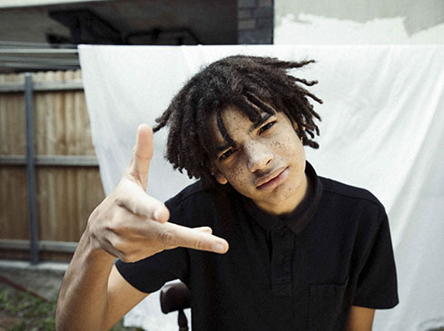 Portrait of a young adult male with dreadlocks and a black polo shirt, posing outdoors with a neutral expression and hand gesture.