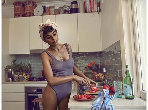 Brunette woman in bodysuit slicing tomatoes in a bright kitchen with white cabinets, brick backsplash, and fruit bowl in natural light.