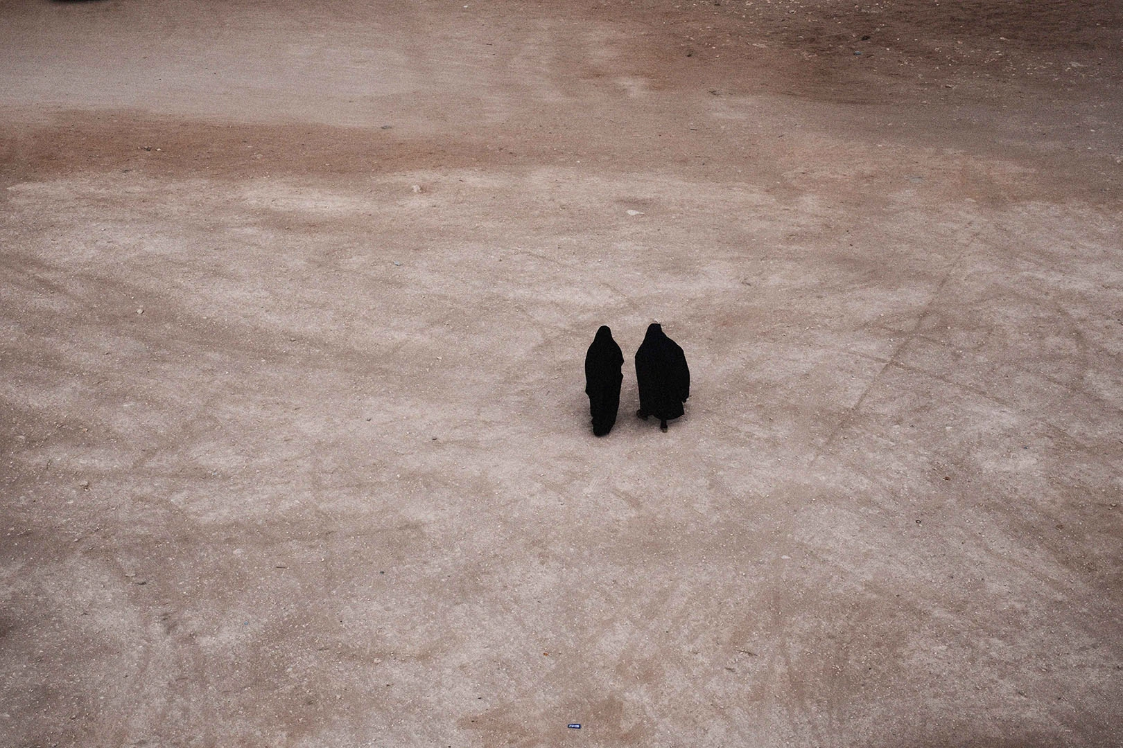 Two figures in black robes walking across an expansive sandy ground viewed from an elevated angle