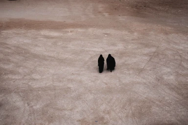 Two figures in black robes walking across an expansive sandy ground viewed from an elevated angle