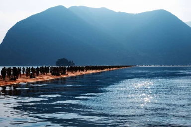 Distant crowd of people walking across a floating orange pier on a body of water with large mountains in the background