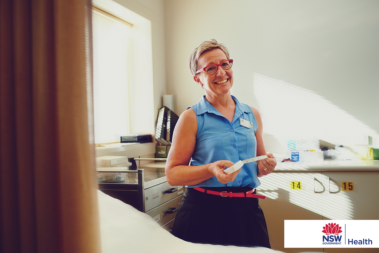 A smiling mature adult female in a blue blouse and glasses stands in a doctor's office, holding a measuring tape and looking at the camera.