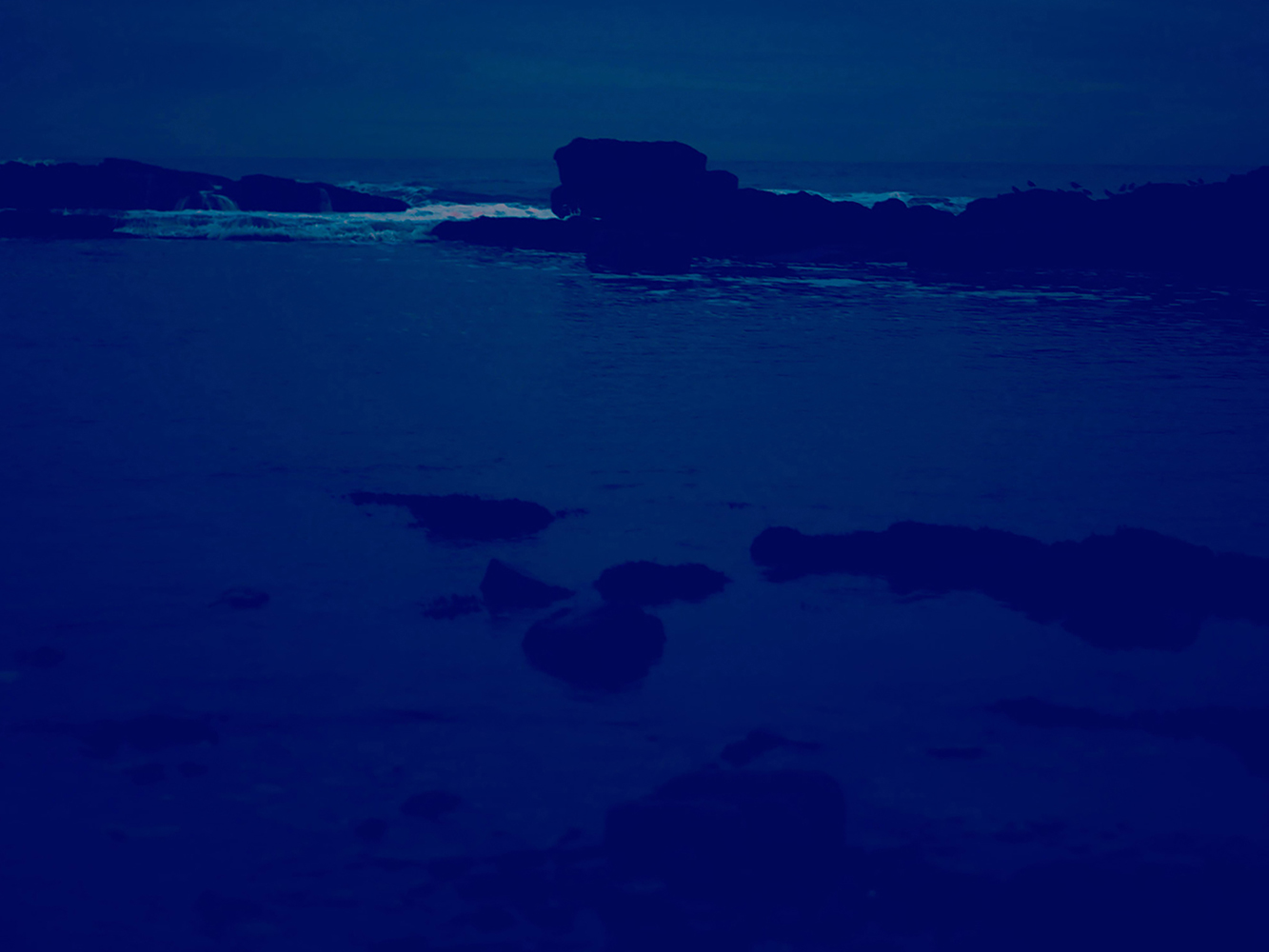 Nighttime seascape with dark water, silhouetted rock formations, and distant whitecaps under a cloudy sky in a monochromatic scene.