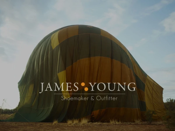 Oversized partially inflated hot air balloon on rural terrain under a sky with light clouds, advertising James Young: Shoemaker & Outfitter.