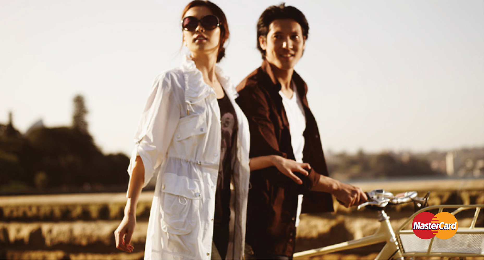 A stylish couple enjoys a bike ride on a sunny day. The woman wears sunglasses and a white jacket, and the man is in a brown shirt.