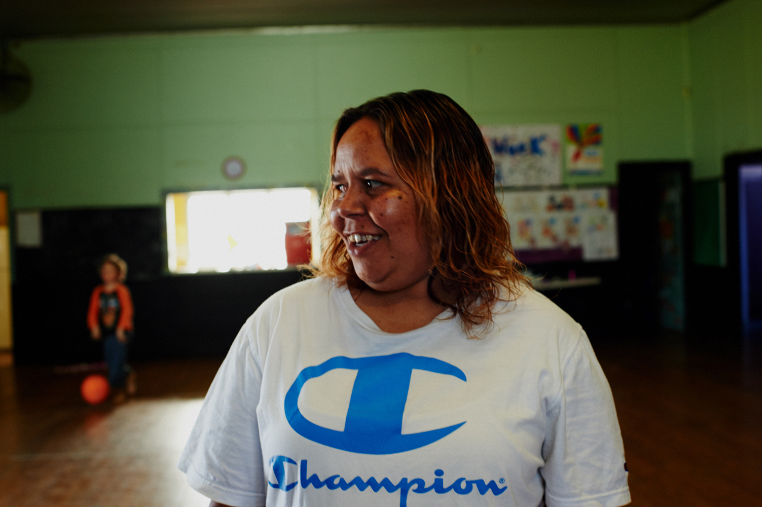 A joyful woman in a Champion T-shirt smiles in a bright indoor space, with a child and ball visible in the background, creating a lively scene.