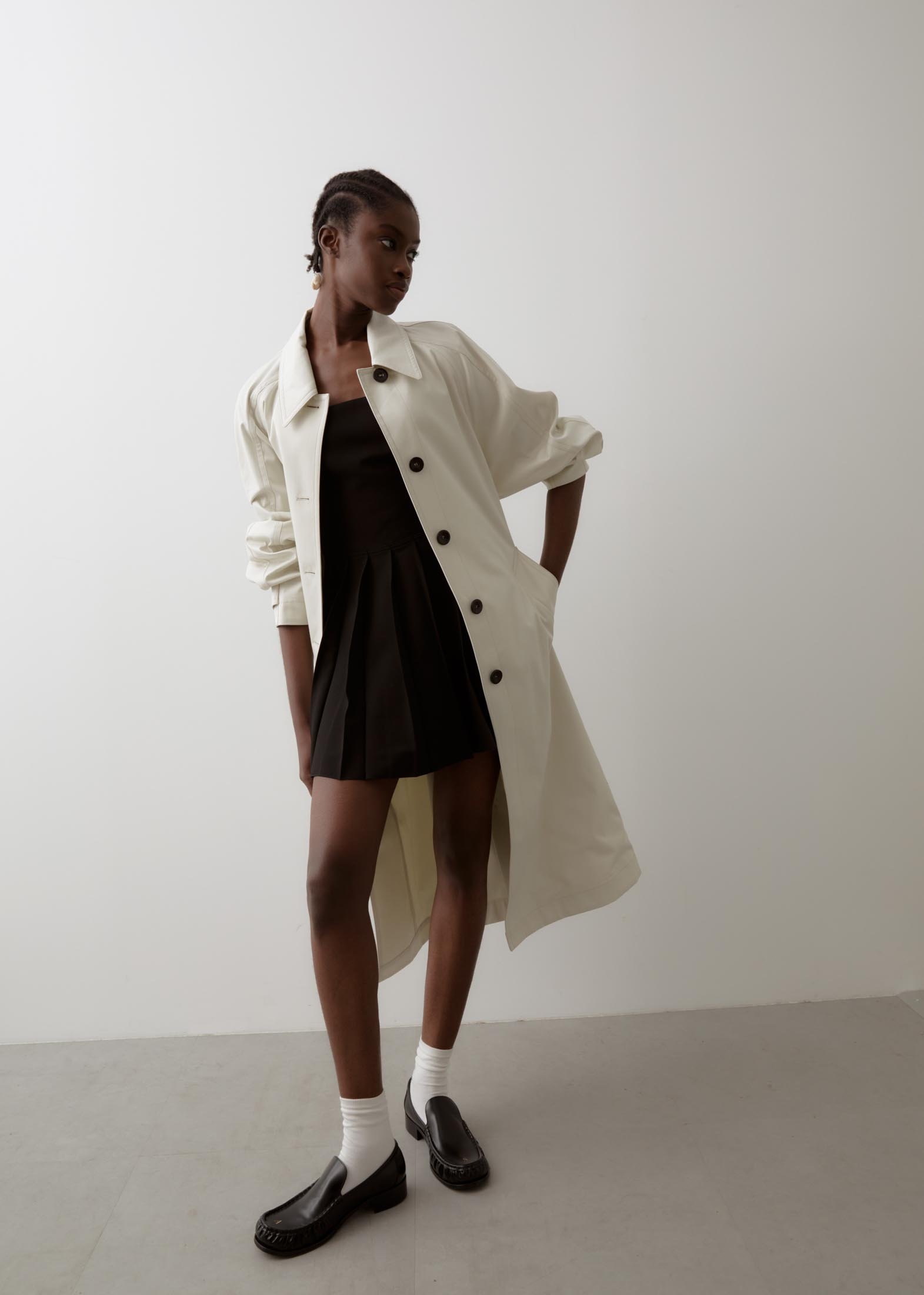 An adult woman with braids poses in a minimalist setting, wearing a black dress, a light trench coat, socks, and black loafers.