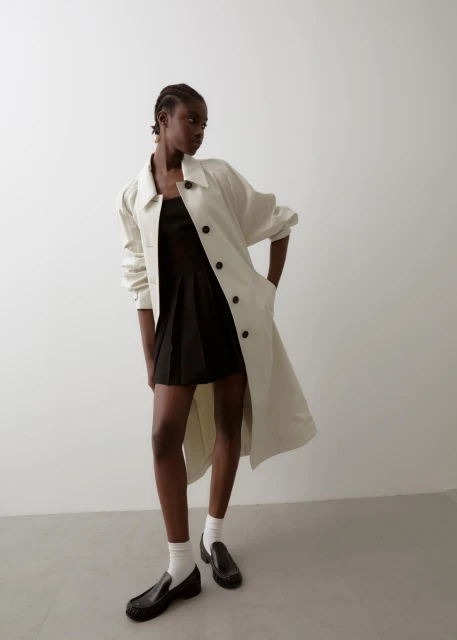 An adult woman with braids poses in a minimalist setting, wearing a black dress, a light trench coat, socks, and black loafers.