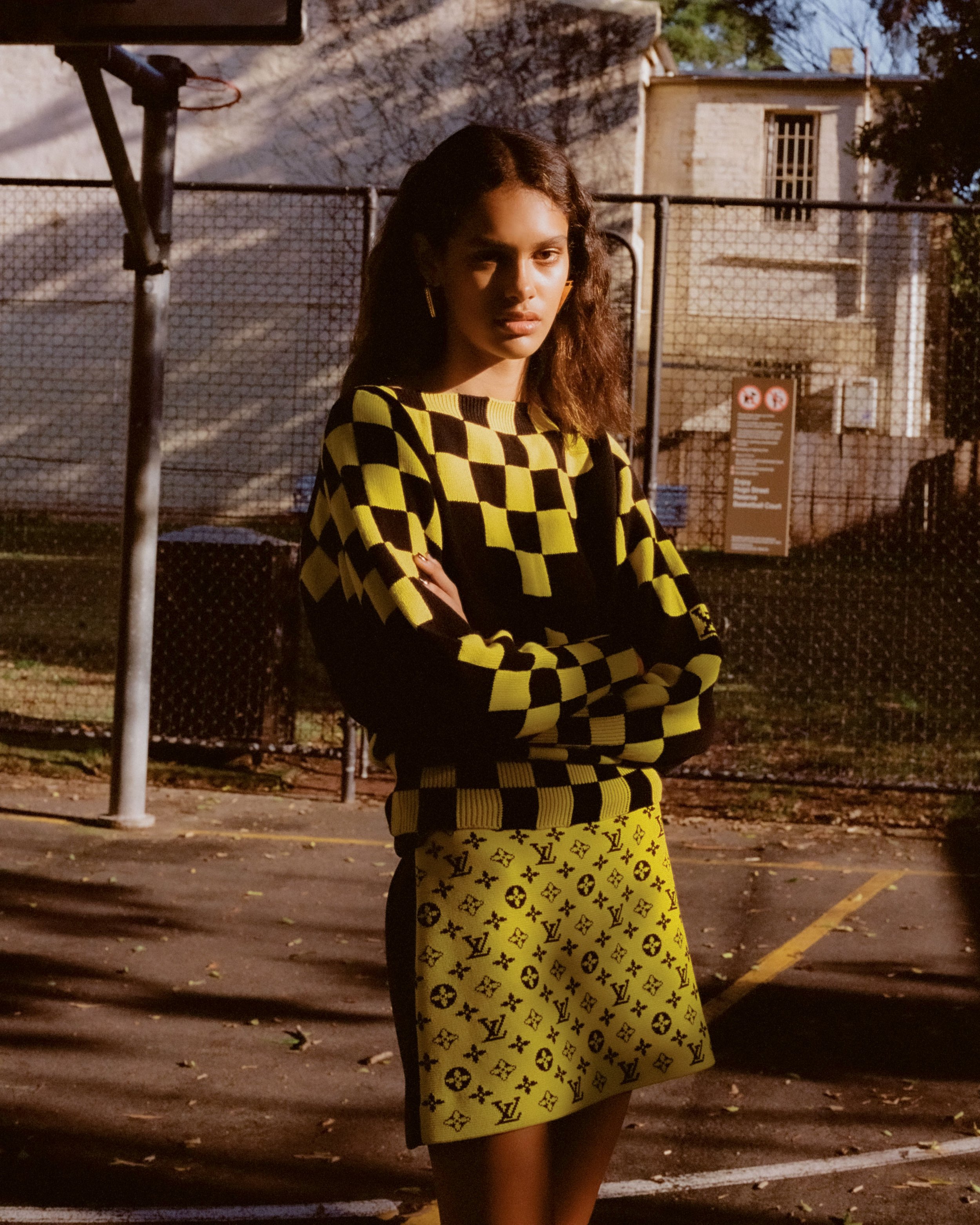 Fashion shot of a young woman wearing a checkerboard sweater and logo skirt, posing on a basketball court with chain-link fence backdrop.