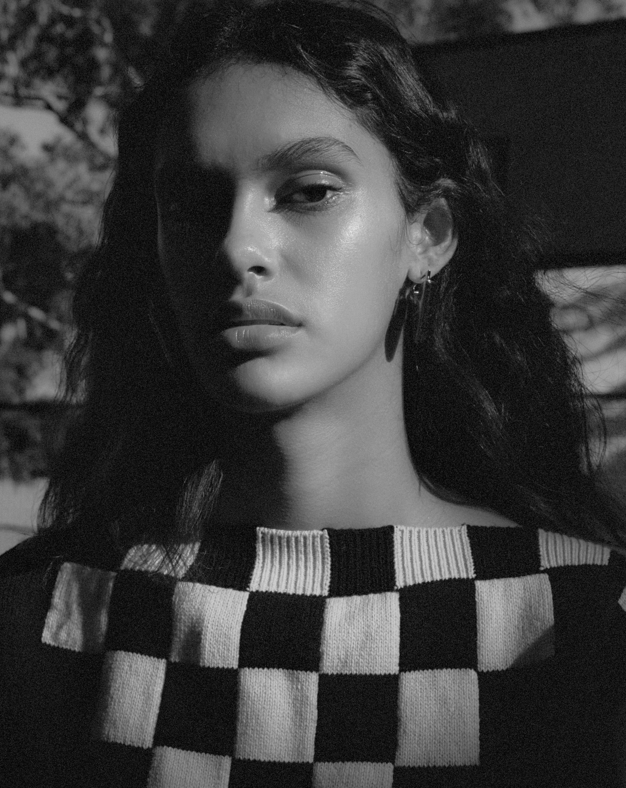 Monochrome portrait of a young woman with dark curly hair wearing a checkered sweater, posing outdoors with diffused natural light.