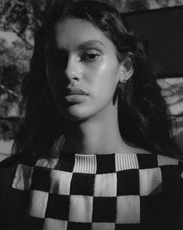 Monochrome portrait of a young woman with dark curly hair wearing a checkered sweater, posing outdoors with diffused natural light.