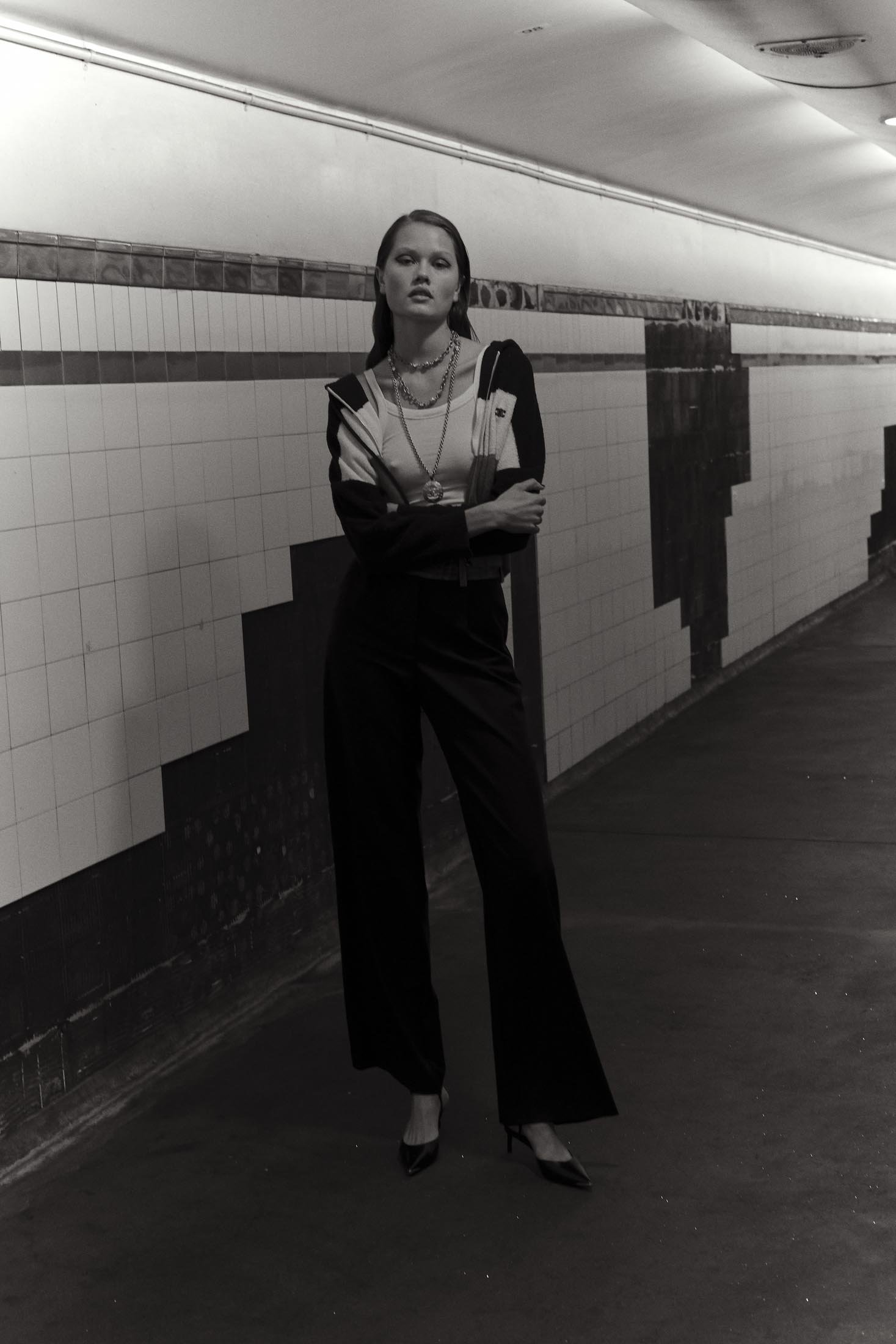 Monochrome fashion shot of a woman posing in a patterned jacket and dark trousers inside a tiled pedestrian tunnel with lighting.