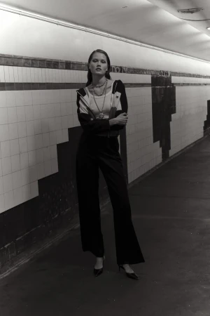 Monochrome fashion shot of a woman posing in a patterned jacket and dark trousers inside a tiled pedestrian tunnel with lighting.