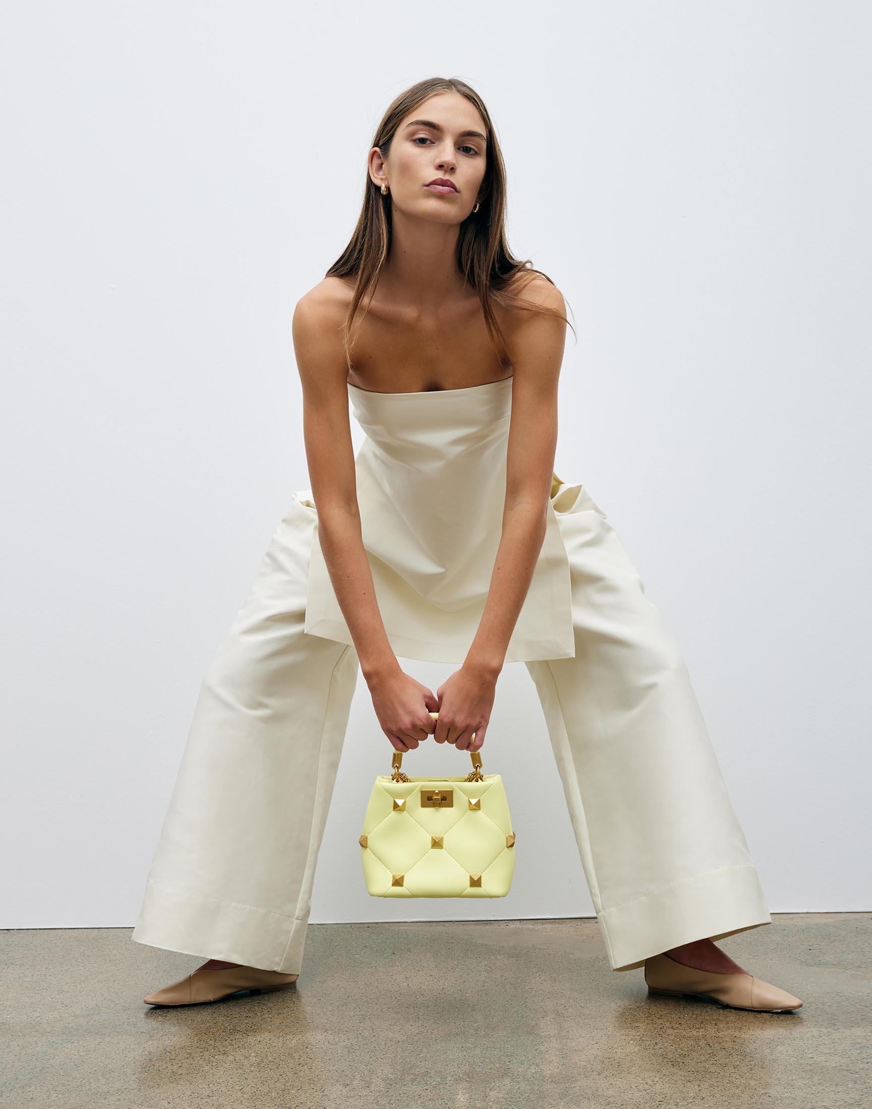 Fashion shoot featuring a woman in a light-yellow strapless top and trousers, holding a light-yellow studded handbag indoors.