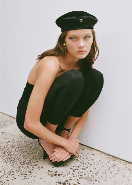Young woman in black beret, tube top and leggings poses in front of white wall with green-blue eyeshadow and black high heels.