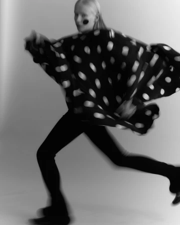 Model in motion wearing a black and white polka dot cape and dark trousers against a light background
