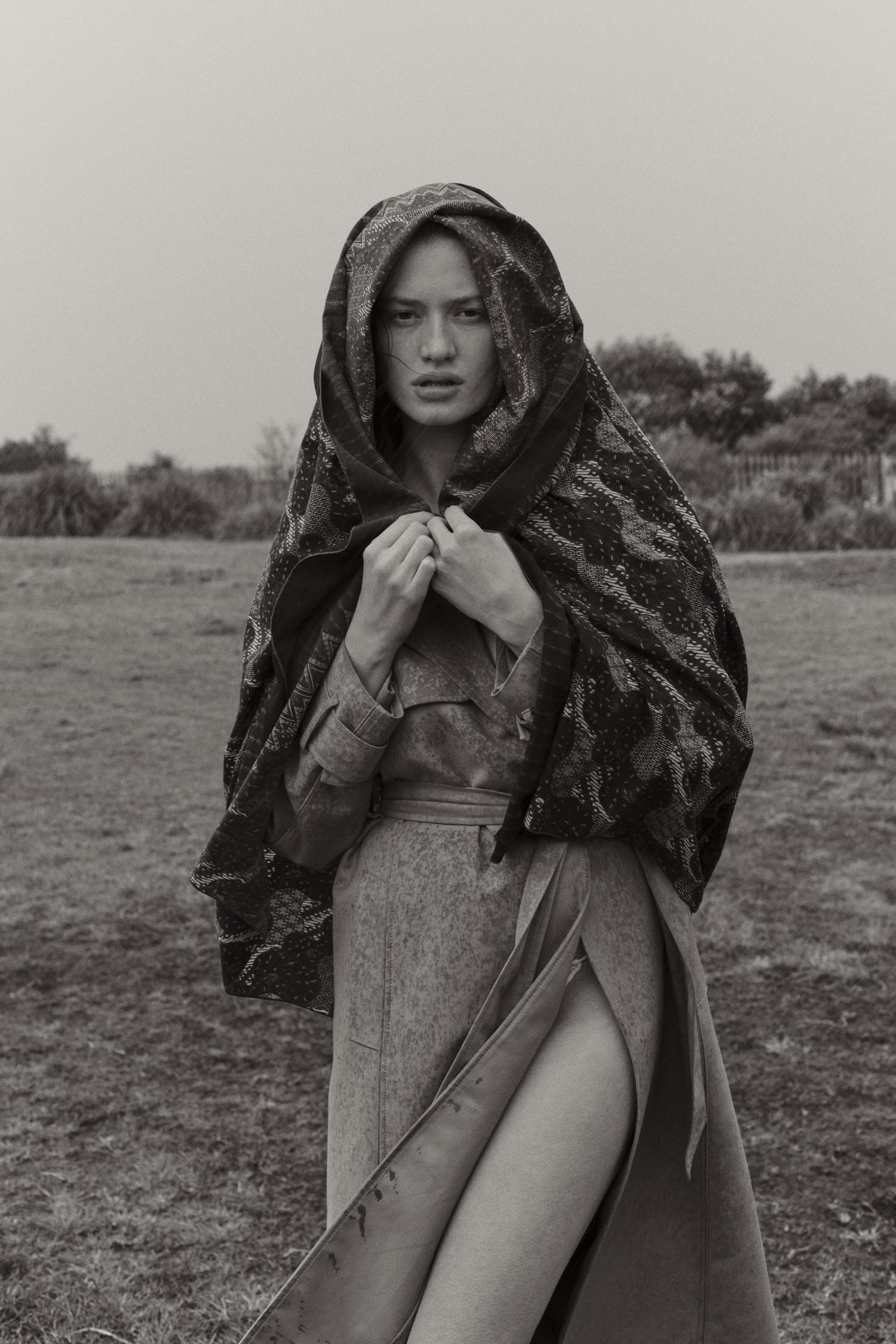 Monochrome portrait of a woman wearing a coat with a wrap, standing in a field with vegetation. The woman is looking at the camera.