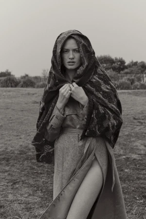 Monochrome portrait of a woman wearing a coat with a wrap, standing in a field with vegetation. The woman is looking at the camera.