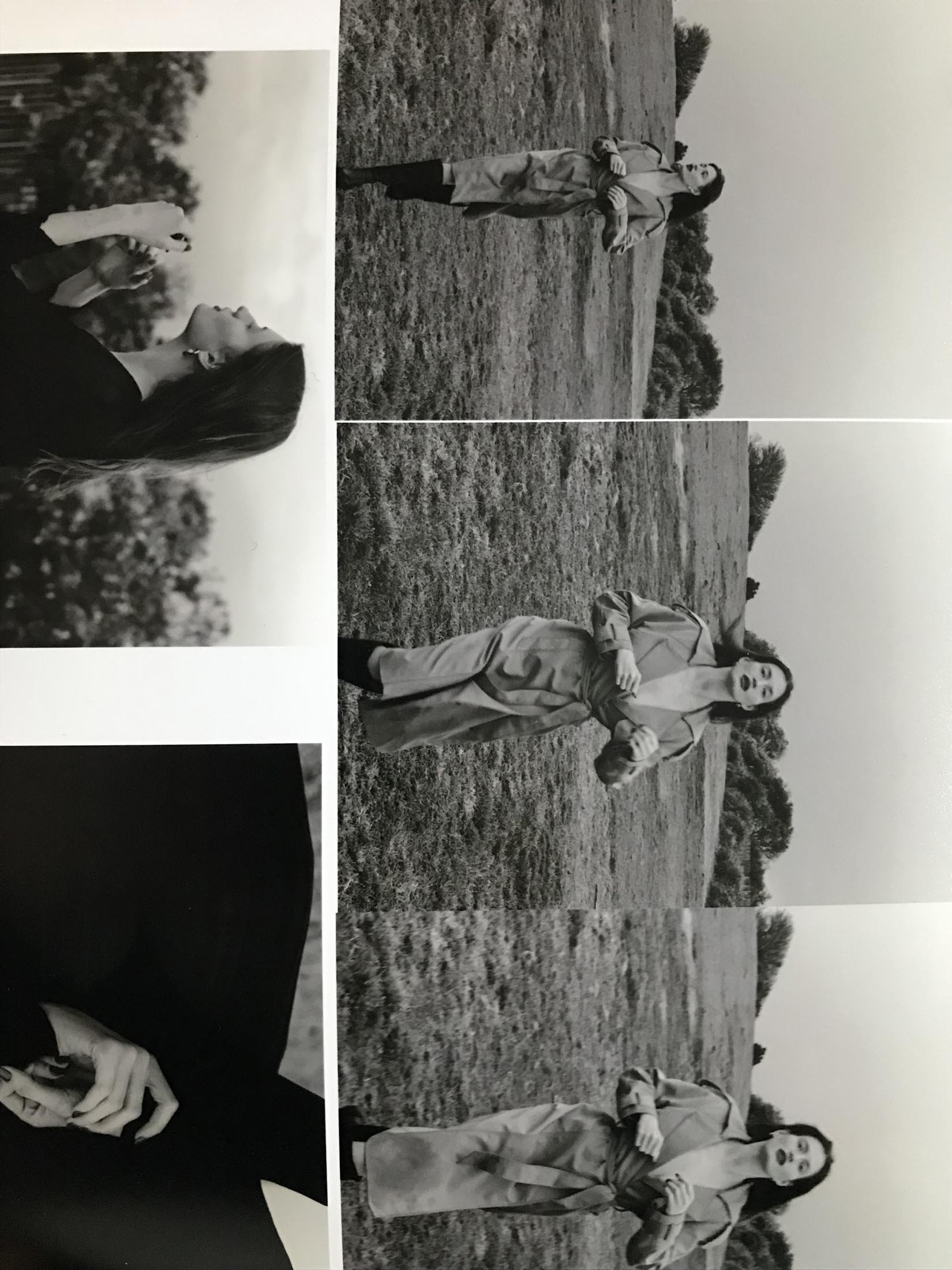 Monochrome fashion shot of a brunette woman in a trench coat, striking a pose in a grassy field, set against a backdrop of trees and sky.