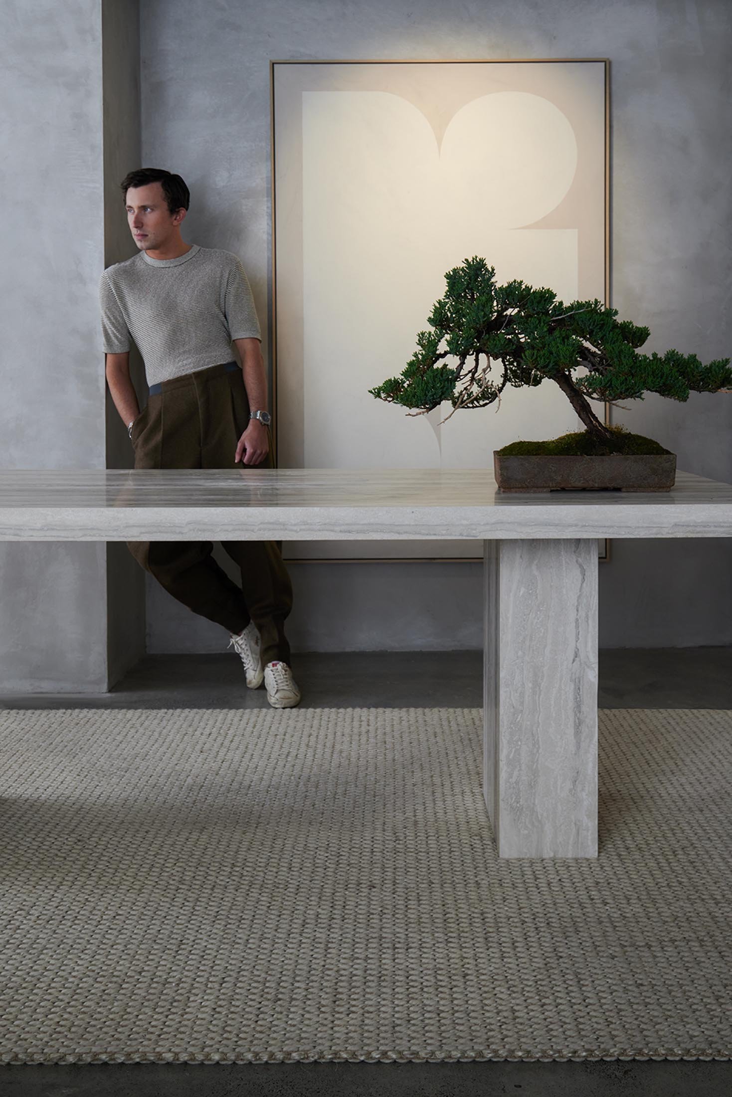 Indoor shot of a man leaning against a wall near a marble table with a bonsai tree and artwork in the background, neutral color palette.