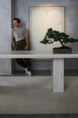 Indoor shot of a man leaning against a wall near a marble table with a bonsai tree and artwork in the background, neutral color palette.