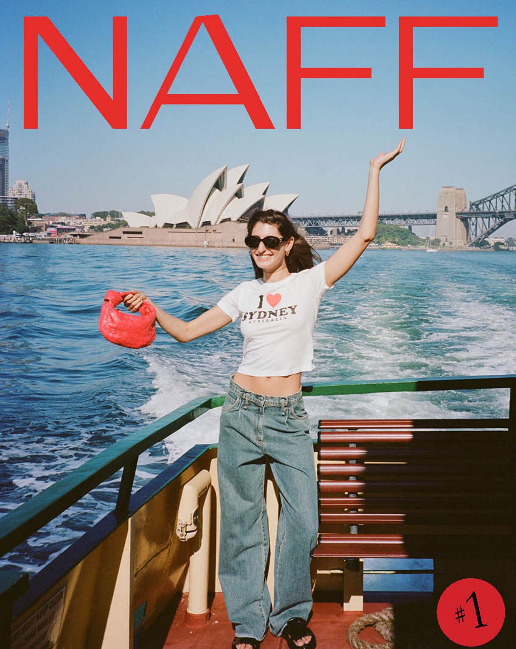 Woman poses on a boat with the Sydney Opera House in the background, wearing jeans and an I Love Sydney t-shirt.