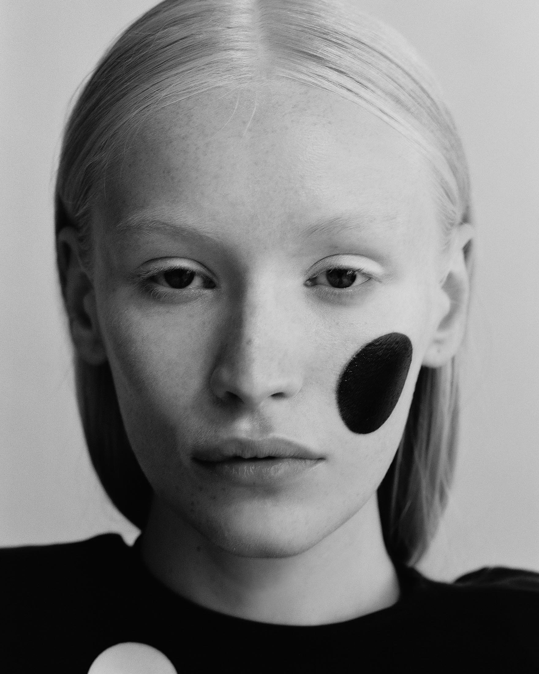 Black and white portrait of a pale-skinned woman with light blonde hair and a black circle of makeup on her right cheek.