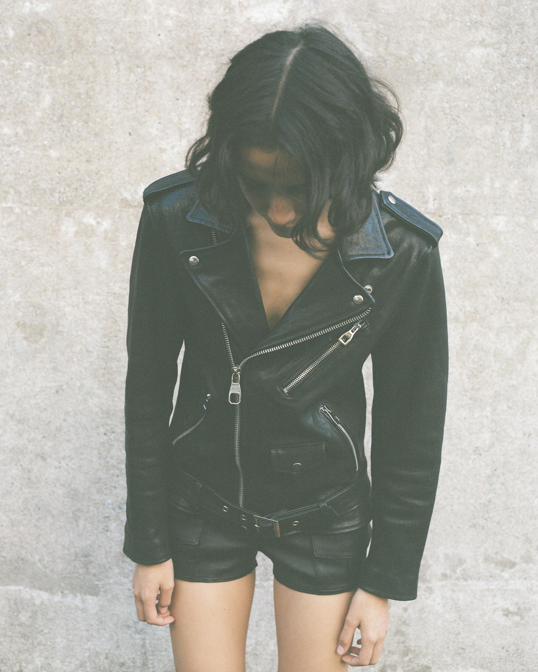 An adult female wearing a black leather jacket and black shorts stands against a grey wall in an indoor or outdoor setting.