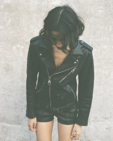 An adult female wearing a black leather jacket and black shorts stands against a grey wall in an indoor or outdoor setting.