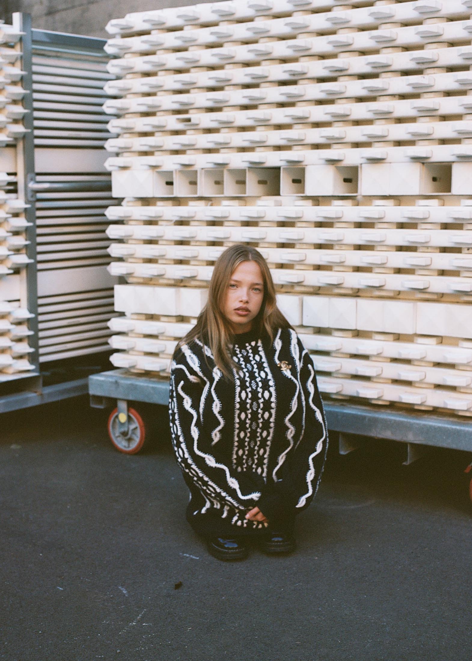 Young woman in oversized sweater squats on grey asphalt in front of stack of white ceramic molds on rolling metal racks outdoors.