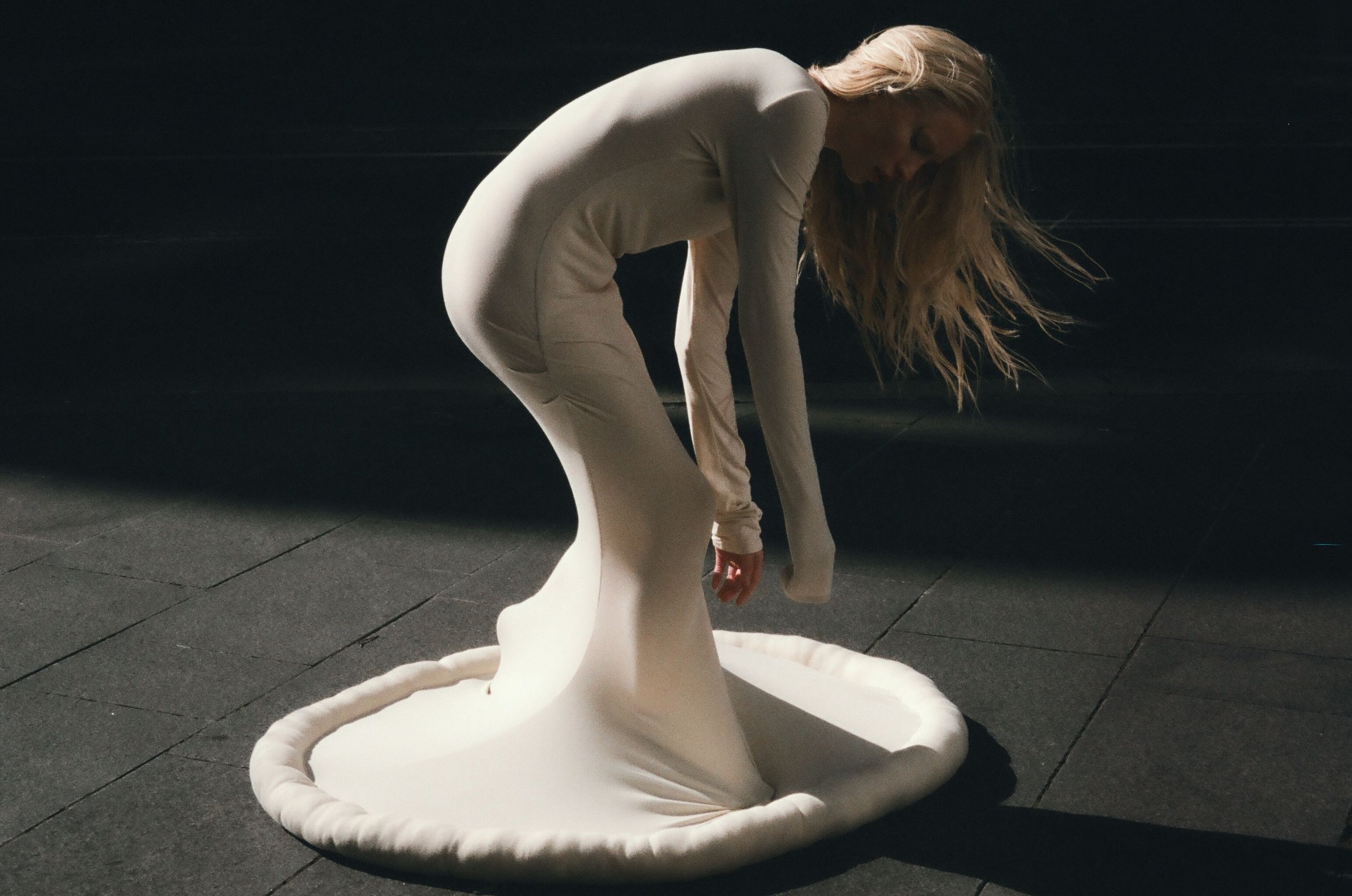 Woman in long white dress bends over in dark studio; blond hair flows down; dramatic, contrast lighting.