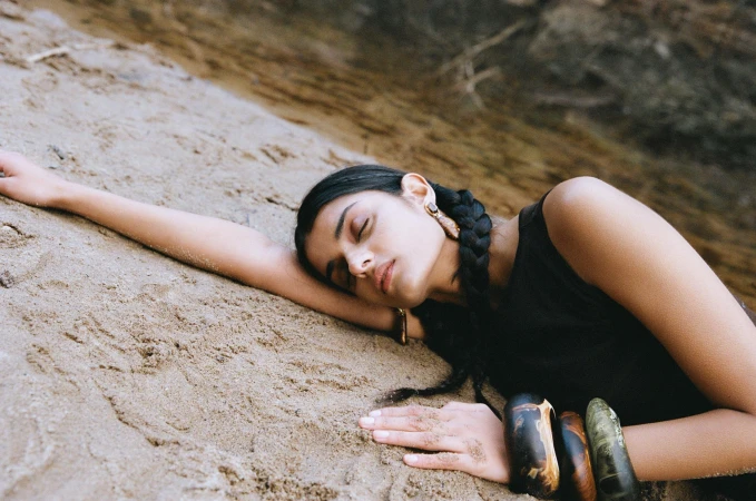Young woman with braided hair and multiple bracelets lies on tan sand near a cliff face in a lifestyle fashion-editorial shot.