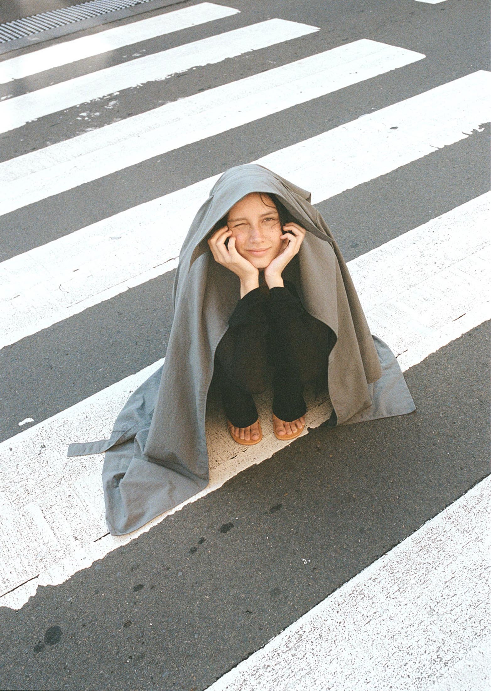 Young adult posing in a crosswalk under a draped gray fabric, resting her chin on her hands, smiling slightly in natural light.