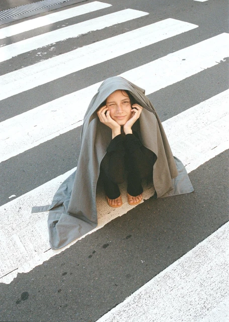 Young adult posing in a crosswalk under a draped gray fabric, resting her chin on her hands, smiling slightly in natural light.