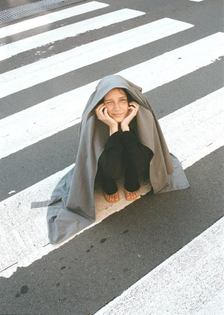 Young adult posing in a crosswalk under a draped gray fabric, resting her chin on her hands, smiling slightly in natural light.