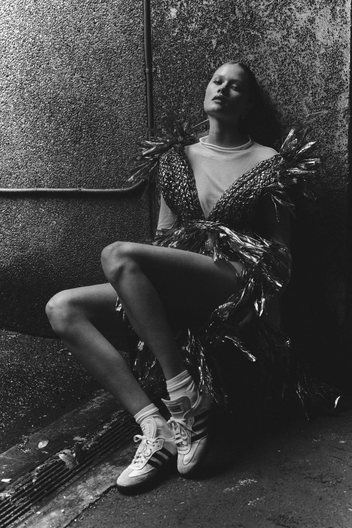 Monochrome portrait of a posed adult female wearing a shiny garment, white t-shirt, white socks, and sneakers while sitting in an urban setting.