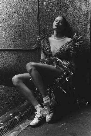 Monochrome portrait of a posed adult female wearing a shiny garment, white t-shirt, white socks, and sneakers while sitting in an urban setting.