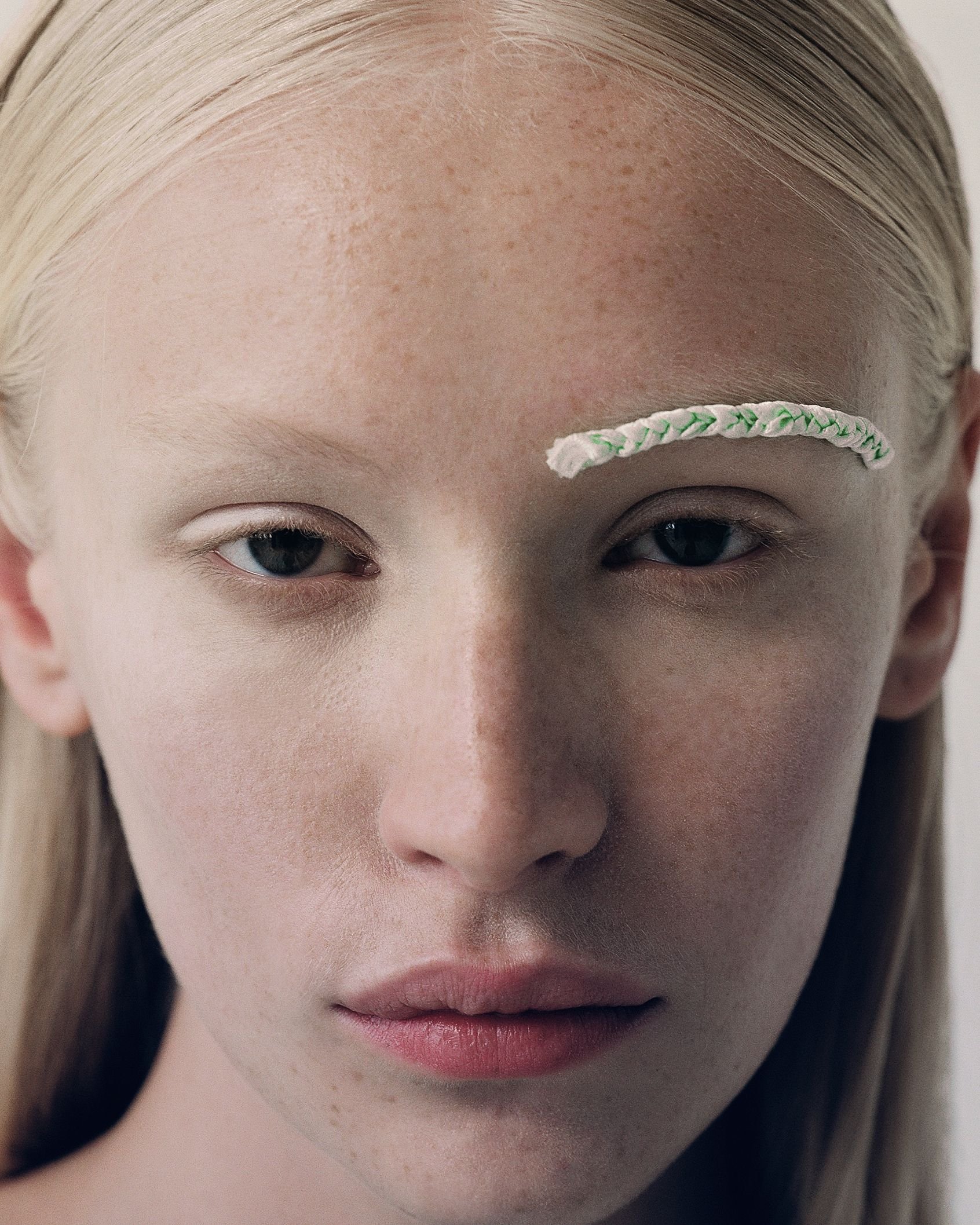 Close-up portrait of a person with light blonde hair and rope on eyebrow, neutral expression, against a plain, soft-lit background.