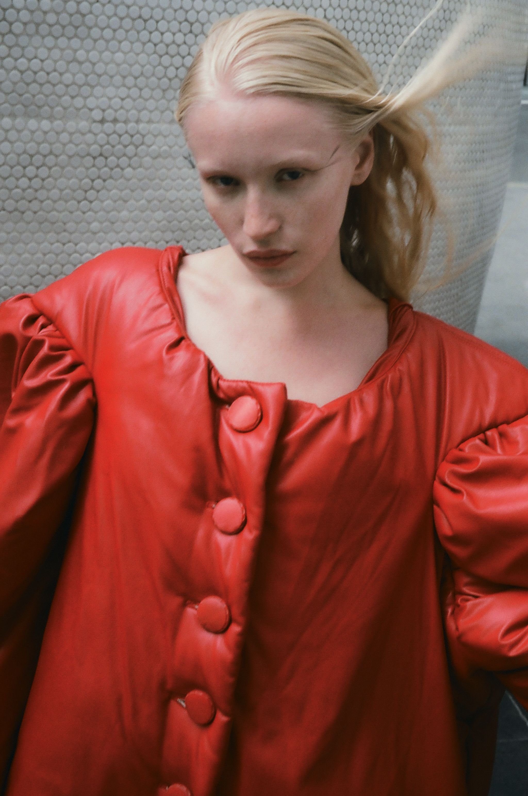 Woman with fair hair wearing a red leather-like puffy jacket posing in front of a textured wall, fashion editorial style.