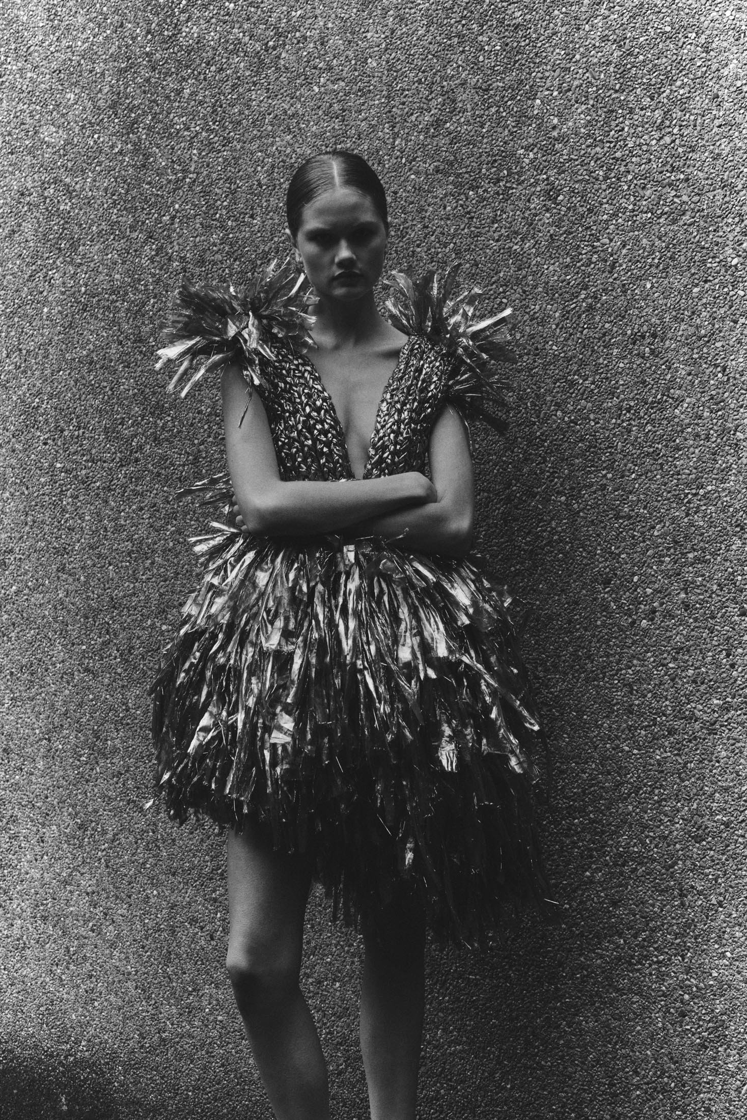 Monochrome fashion portrait of a woman in an avant-garde dress with feathery textures, posing against a textured gray backdrop.