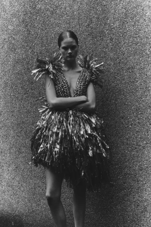 Monochrome fashion portrait of a woman in an avant-garde dress with feathery textures, posing against a textured gray backdrop.