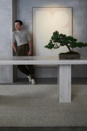 Man leaning against a concrete wall beside a large travertine dining table featuring a potted bonsai tree
