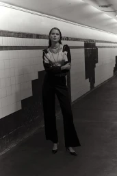 Model standing in a tiled underpass with arms crossed, wearing a dark jacket, trousers, and layered necklaces.