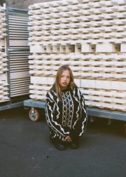 A young woman wearing a patterned black and white knit sweater kneeling in front of stacked building materials.