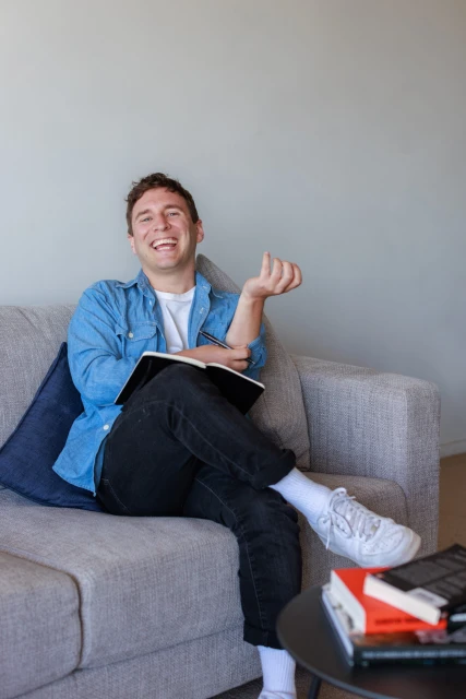 Smiling man in a blue shirt sits on a gray couch, holding a notebook and pen, with books stacked on a side table in a bright indoor setting.