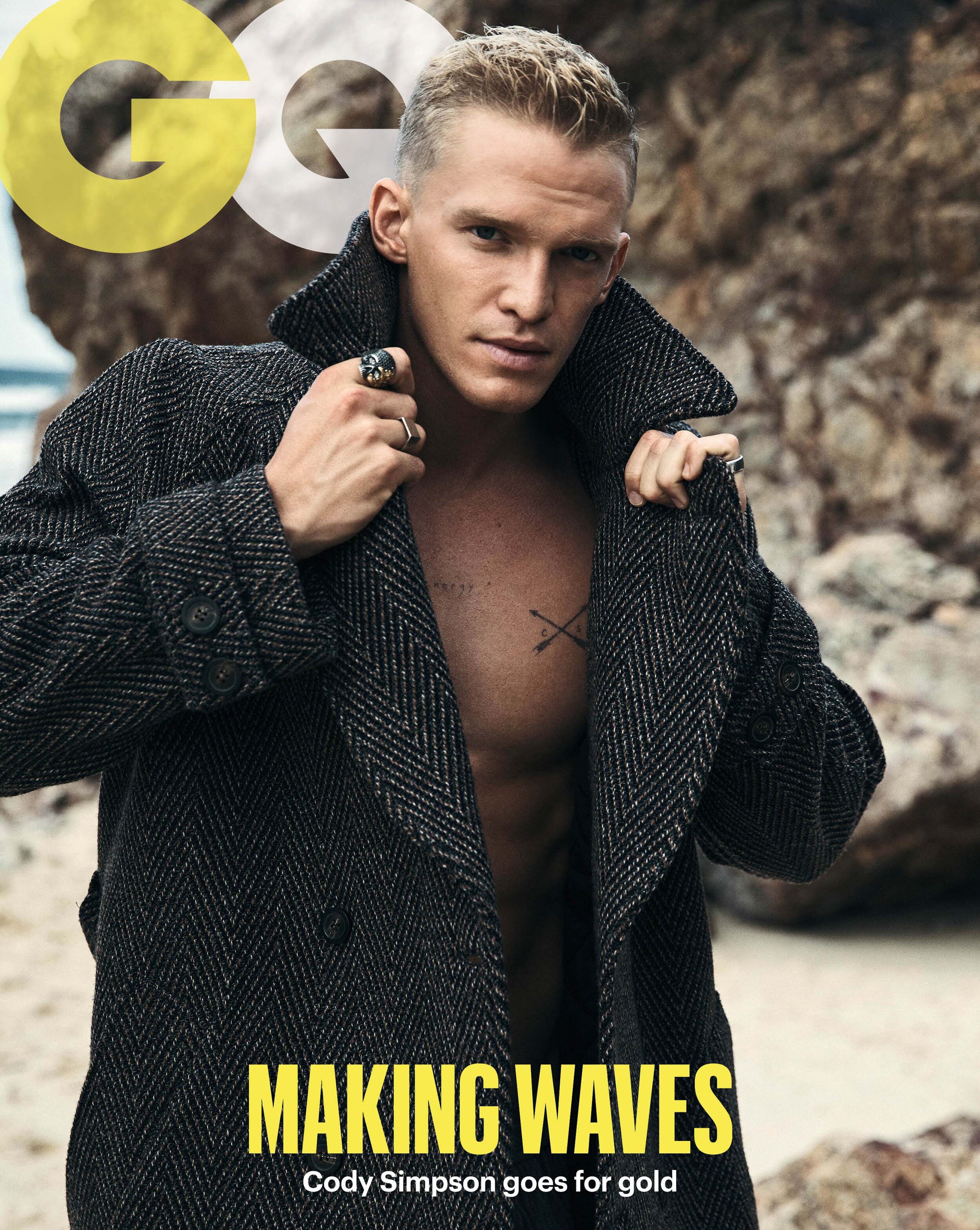 Portrait of Cody Simpson on a beach, wearing an open dark herringbone coat, showcasing his torso with a tattoo. Magazine cover.